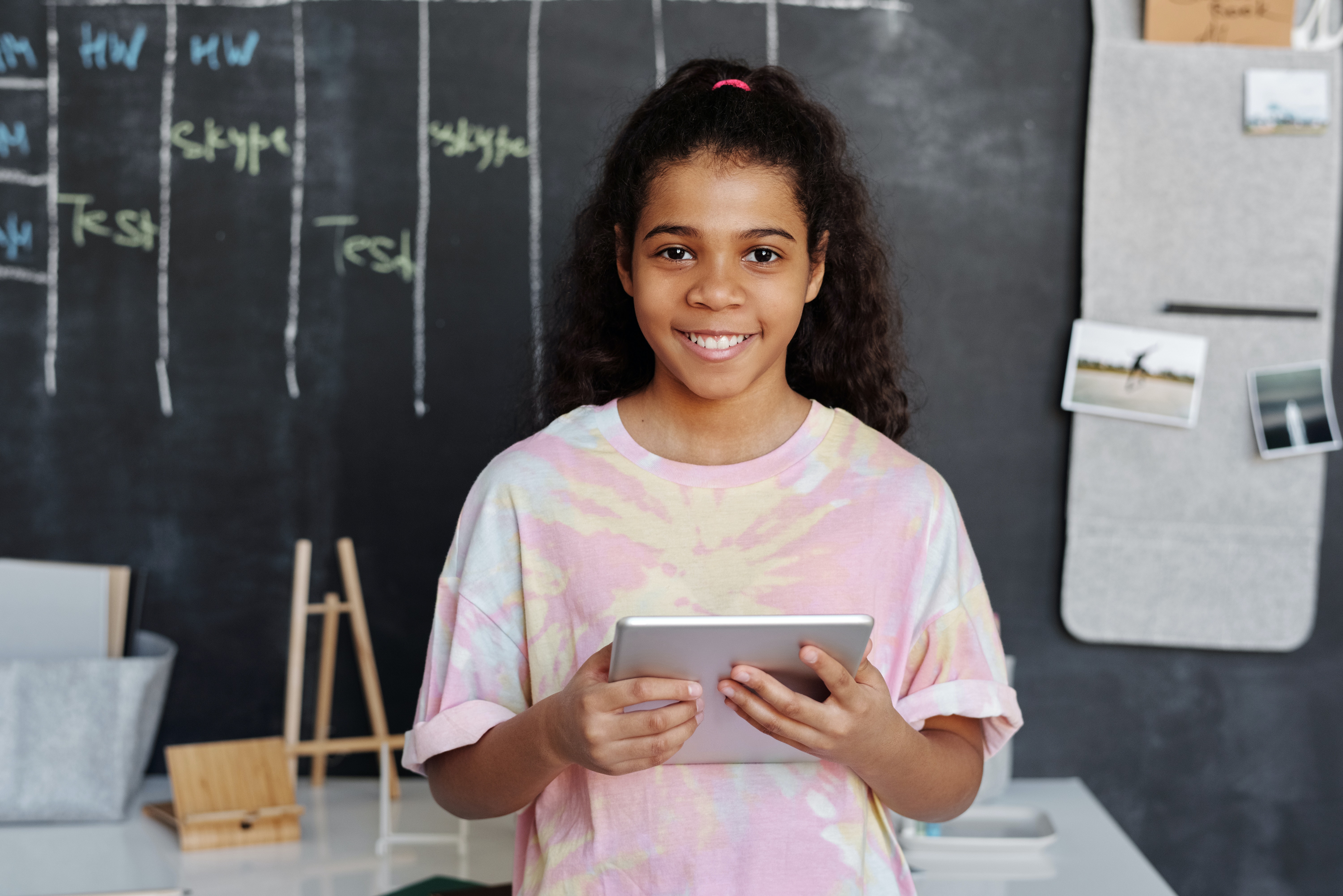 A young girl in a pink shirt holds a tablet in her classroom.